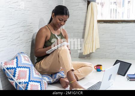 Porträt eines brünetten Mädchens, das sich Notizen machte, auf dem Boden saß, mit Laptop und Kaffee am Fenster Stockfoto