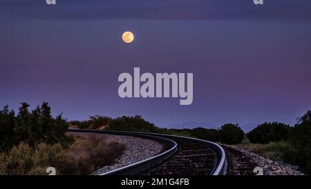Der Vollmond im violett-blauen Sonnenaufgang über gewundenen Bahngleisen auf dem Land Stockfoto