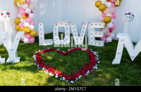 Herzform aus Rosenpedalen mit LIEBESZEICHEN für einen Heiratsantrag in einem Garten auf Gras Stockfoto
