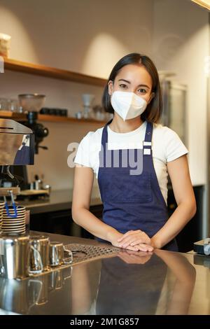 Vertikale Aufnahme eines freundlichen Baristas, weibliche Mitarbeiterin in einer medizinischen Maske, arbeitet hinter dem Tresen mit Kunden, serviert Kaffee im Café, steht hinein Stockfoto
