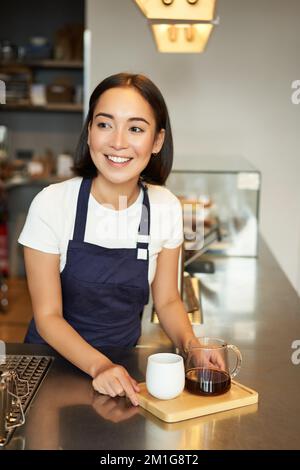 Vertikale Aufnahme einer lächelnden Barista, die Kaffee serviert, eine Portion Kaffee zubereitet, für den Gast im Café filtert und hinter der Theke eine blaue Schürze trägt Stockfoto