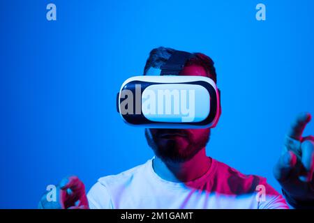 Ein junger Mann, der eine Virtual-Reality-Brille trägt und mit einem Freund im Metaversum spaziert. Ein bärtiger Mann, der Virtual-Reality-Videospiele in einem Studio ausprobiert. Stockfoto