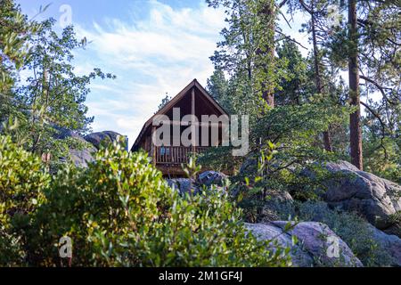 Blick auf die Landschaft auf ein Holzhaus, umgeben von großen Felsen und grünen Bäumen unter blauem Himmel Stockfoto