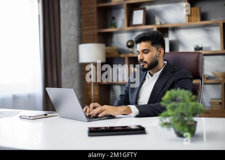 Junger, bärtiger Geschäftsmann, der während der Arbeit im Büro auf einem Laptop tippt. Konzentrierter Erwachsener, erfolgreicher Mann in offiziellem Anzug, der drinnen am Holzschreibtisch sitzt. Stockfoto
