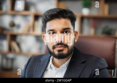 Nahaufnahme eines bärtigen, gutaussehenden, erfolgreichen, lächelnden Geschäftsmanns aus dem Nahen Osten, der an seinem Stuhl sitzt und in seinem modernen Büro arbeitet. Stockfoto