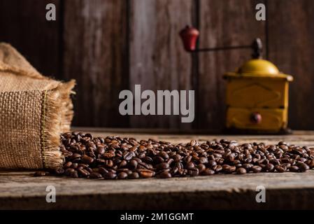 Geröstete Kaffeebohnen, die aus einem Sack auf einem Holztisch kommen und im Hintergrund eine alte Kaffeemühle. Stockfoto