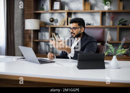 Selbstbewusster Geschäftsmann mit Anzug und Brille, der während eines Video-Chats am Schreibtisch mit einem Laptop sitzt, fokussierter ernster Mann, der mit einem neuen Projekt arbeitet, Schüler, die online studieren, Forschungsarbeit Stockfoto