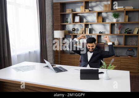 Aufgeregt schreiender junger, bärtiger Geschäftsmann oder Unternehmer, der mit einem Laptop und Tablet arbeitet und mit erhobenen Händen triumphiert, weil er ein erfolgreiches Geschäft abgeschlossen hat. Stockfoto