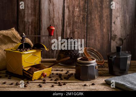 Alte Kaffeemühle mit Bohnen und frisch gemahlenem Kaffee, neben einer offenen italienischen Kaffeemaschine, bereit für die Kaffeezubereitung, im Hintergrund eine Kanne mit Röstung Stockfoto