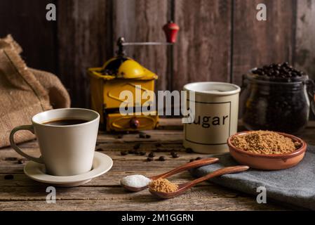 Eine Tasse frisch zubereiteter Kaffee neben einigen Holzlöffeln mit Weißzucker und eine andere mit braunem Zucker und den Gläsern dahinter, im Hintergrund ein alter Kaffee Stockfoto