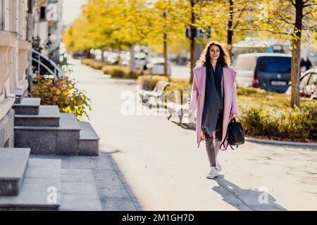 Frau Herbststadt. Eine Frau in einem rosa Kunstpelzmantel, die im Herbst an einem sonnigen Tag auf einer Straße posiert. Bäume mit gelben Blättern entlang der Straße. Stockfoto