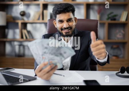 Portrait eines glücklichen jungen, attraktiven männlichen Buchhalters, der Bargeld in der Hand hält, Geld in einem modernen Büro zählt und Daumen nach oben zeigt. Konzept der Finanzplanung und Mittelzuweisung. Stockfoto
