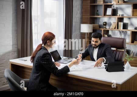 Ein gutaussehender, bärtiger Geschäftsmann, der seiner rothaarigen Geschäftsfrau Papiere mit neuer Vereinbarung auf einem Desktop zeigt, sitzt am Tisch in einem modernen Büro. Stockfoto