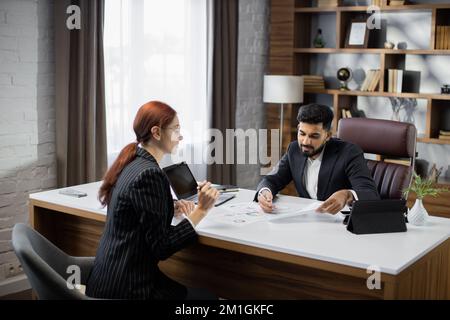 Ein gutaussehender, bärtiger Geschäftsmann, der seiner rothaarigen Geschäftsfrau Papiere mit neuer Vereinbarung auf einem Desktop zeigt, sitzt am Tisch in einem modernen Büro. Stockfoto