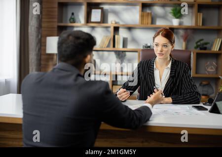Eine rothaarige Geschäftsfrau, die ihrem gutaussehenden, bärtigen Geschäftsmann Papiere mit neuer Vereinbarung auf einem Schreibtisch zeigt, sitzt am Tisch in einem modernen Büro. Stockfoto