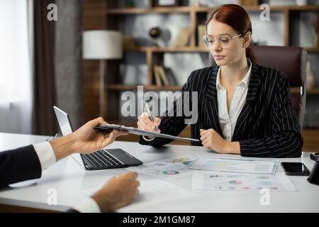 Fokussierte junge Geschäftsfrau, die einen Vertrag mit einem erfahrenen Anwalt unterschreibt. Konzentrierte Finanzberaterin, die bei einem Treffen im Büro gegenüber einer Kundin einen Platz zur Unterzeichnung auf einem Vertragsdokument in Papierform vorweist. Stockfoto