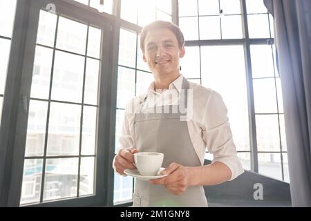 Ein Kellner in einer grauen Schürze hält eine Tasse Kaffee. Der Barista gibt eine Tasse heißen Kaffee in einem Café vor dem Hintergrund eines großen Fensters. Hochwertiges Foto Stockfoto