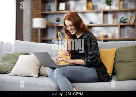 Junge Frau, die für ihr Unternehmen recherchiert. Lächelnde Frau, die auf dem Sofa sitzt und sich beim Surfen im Internet entspannt. Glückliches Mädchen, das zu Hause in der Freizeit im Internet surft. Stockfoto