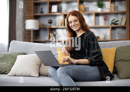 Junge Frau, die für ihr Unternehmen recherchiert. Lächelnde Frau, die auf dem Sofa sitzt und sich beim Surfen im Internet entspannt. Glückliches Mädchen, das zu Hause in der Freizeit im Internet surft. Stockfoto