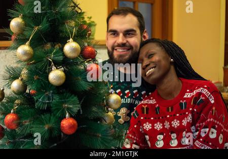 Gemischtrassiges Paar, das sich an weihnachten vor einem dekorierten Baum umarmt, während es weihnachtspullover trägt. Stockfoto