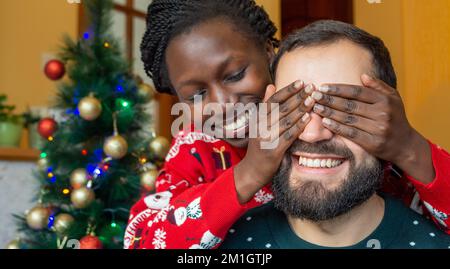 Glückliche afrikanische Frau, die die Augen eines bärtigen Mannes mit ihren Händen bedeckt, glücklich, ein weihnachtsgeschenk vor dem weihnachtsbaum zu bekommen, in einem weihnachtsswea Stockfoto