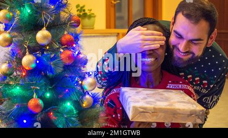 Glücklicher Mann, der die Augen einer jungen afrikanerin mit seinen Händen bedeckt, glücklich, ein weihnachtsgeschenk vor dem weihnachtsbaum zu erhalten, das Weihnachtsschweiß trägt Stockfoto