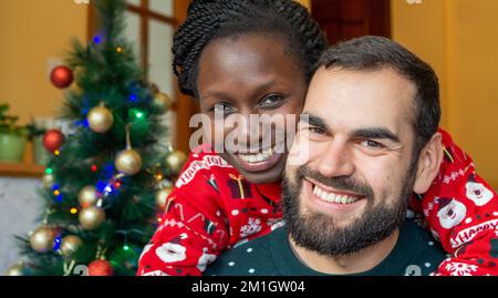 Gemischtrassiges Paar, das sich an weihnachten vor einem dekorierten Baum umarmt und in die Kamera schaut, während es weihnachtspullover trägt. Stockfoto