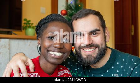 Gemischtrassiges Paar, das sich an weihnachten vor einem dekorierten Baum umarmt und in die Kamera schaut, während es weihnachtspullover trägt. Stockfoto