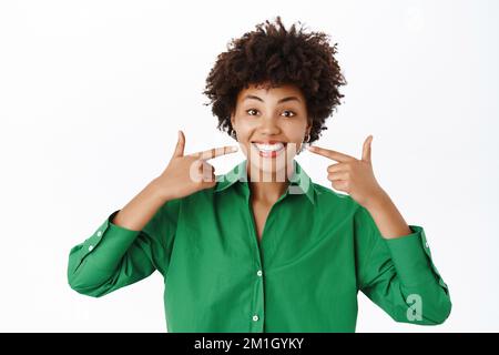 Zahnärztliche Versorgung. Ein lächelndes afroamerikanisches Mädchen, das auf seine weißen, perfekten Zähne zeigt, ein Lächeln ohne Zahnspange oder Zahnhalter zeigt, weißer Hintergrund Stockfoto