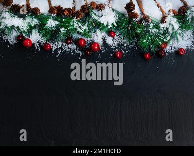 Weihnachtlicher Schnee, Tannenspitzen und rote Beeren auf dunklem Steinhintergrund Stockfoto