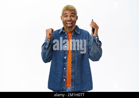 Aufgeregter, lächelnder Schwarzer, der mit dem Handy tanzt, vor Begeisterung und Glück springt, auf dem Smartphone gewinnt, weißer Hintergrund Stockfoto
