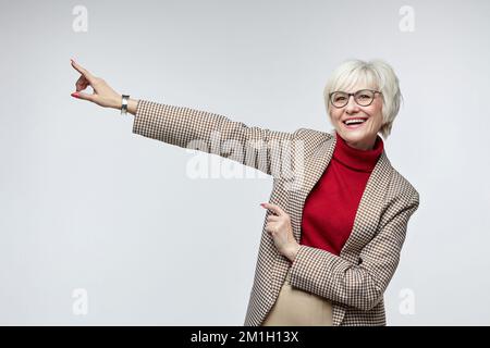 Schöne ältere Frau mit Brille zeigt mit dem Zeigefinger auf einen leeren Sitz. Werbekonzept einer Geschäftsfrauenführerin. Stockfoto
