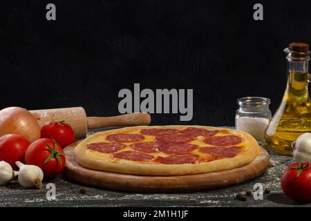 Fertige Pizza mit Salami auf einem Holzbrett und schwarzem Hintergrund. Kulinarischer Hintergrund. Stockfoto