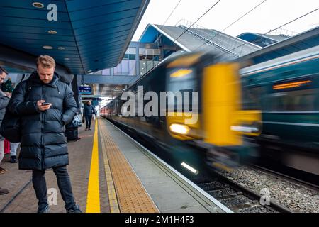 Züge, die in einem Bewegungsunschärfe gesehen werden, wenn einer ankommt und ein anderer vom Bahnhof Reading in Großbritannien abfährt, während ein Mann sein Telefon benutzt und wartet. Stockfoto