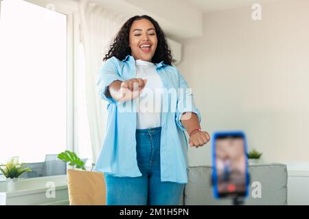 Fröhliche, schwarze, dicke Frau, die in der Handykamera tanzt und zu Hause Videos mit dem Telefon auf dem Stativ filmt Stockfoto