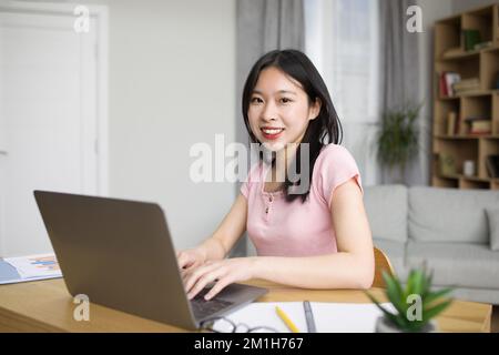 Porträt einer glücklichen asiatischen Frau, die einen Laptop benutzt, zu Hause am Schreibtisch sitzt und vor der Kamera lächelt, Kopierraum Stockfoto