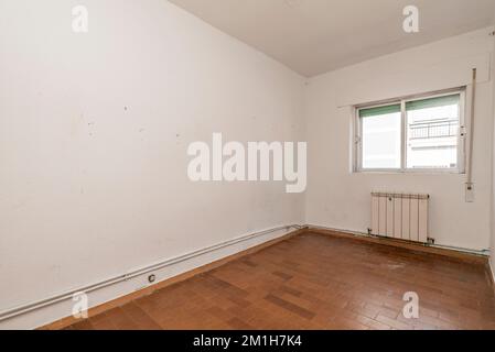 Leeres Wohnzimmer mit hässlich braunen Fliesenböden, freiliegenden Heizrohren und weißen Aluminiumheizkörpern Stockfoto