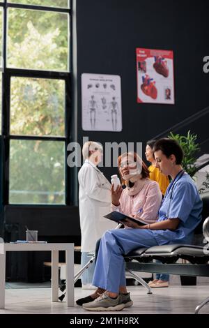 Arzthelferin, die verletzten Patienten während eines Arzttermins Flaschentabletten gibt und die medikamentöse Behandlung erklärt. Seniorin trägt Halskragen und konsultiert in der Krankenhauslobby Stockfoto