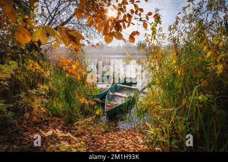 Am frühen Morgen auf dem Herbstfluss erleuchtet die Sonne die halb untergetauchten Boote, die am Ufer festgemacht sind Stockfoto