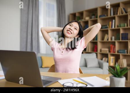 Entspannte asiatische Studentin, die mit den Händen hinter dem Kopf neben dem Laptop sitzt, eine Pause nach dem Online-Kurs hat, Kopierbereich Stockfoto