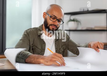 Seriöser männlicher Brilleningenieur, der mit Projektzeichnungen am Arbeitsplatz im Innenbereich des Heimbüros arbeitet Stockfoto