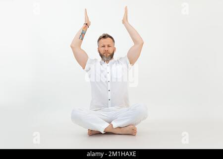 Konzentrierter, sportlicher Mann mittleren Alters mit Bart, der mit gekreuzten Beinen auf dem Boden sitzt und die Hände auf weißem Hintergrund hebt. Yoga. Stockfoto