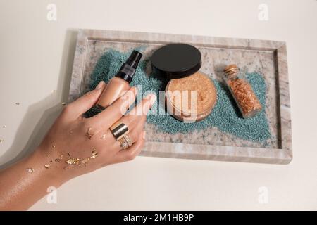 Gekürzte Frauenhand mit Ringen, goldene Glitzer halten die Fundamente der Tönungscreme über dem Tablett mit Pulver und Granulat Badesalz Stockfoto