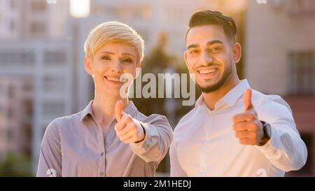 Die beiden multiethnischen Geschäftspartner, die kaukasische Frau und der hispanische Mann, stellten den Daumen hoch und standen bei Sonnenuntergang im Freien. Multirassisches Paar Stockfoto