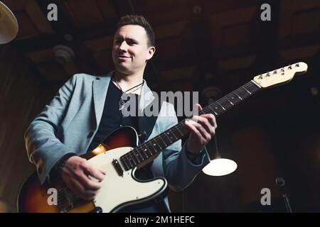 Ein Musiker, der auf einer Gitarre spielt. Weißer Erwachsener in einer Rockband, der Gitarre spielt. Bühnenbeleuchtung. Innenaufnahme mit niedrigem Winkel. Hochwertiges Foto Stockfoto