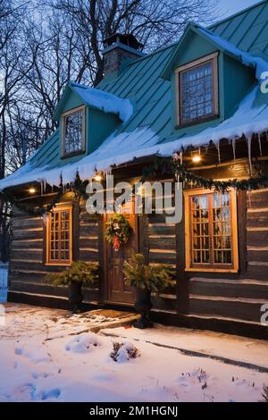 Altes zweistöckiges Blockhaus im Canadiana Landhausstil aus dem Jahr 1800er mit Weihnachtsdekorationen, die im Winter in der Abenddämmerung beleuchtet werden. Stockfoto