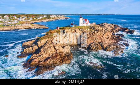 Wellen stürzen über Felsen über Maine Island mit Leuchtturm und Blick auf Häuser auf dem Festland Stockfoto