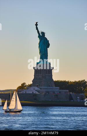 Ein kleines Segelboot fährt bei Sonnenuntergang an der Silhouette der Freiheitsstatue in New York City vorbei Stockfoto