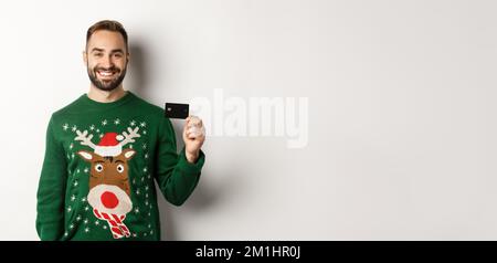 Weihnachtsferien und Shopping-Konzept. Junger Mann im Winterpullover mit Kreditkarte, auf weißem Hintergrund stehend Stockfoto