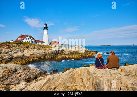 Ein Paar genießt den Blick auf den Leuchtturm an der Küste von Maine, der auf Felsen sitzt Stockfoto